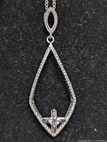 Close-up photo of silver pendant on black background showing teardrop outline with embedded create stones and cross centerpiece with larger clear stone.