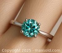 Front view of the silver ring worn on a finger, displaying the 1.1ct round blue-green moissanite gemstone in a four-prong setting.