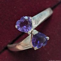 Close-up photo of a silver ring with two faceted purple amethyst stones, one heart shaped and one pear shaped, on an open band design placed against a dark red background.