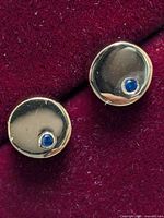 Close-up photo of one round sterling silver earring with a small blue sapphire stone set in it, shown on a red fabric background.