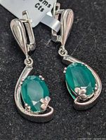 Close-up front and side views of pair of sterling silver drop earrings each set with one oval emerald gemstone in prong settings with floral silver accents.