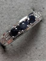 Close-up photo of sterling silver ring with three blue sapphires in center and clear stones on sides on a grey background.