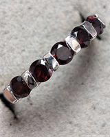 Close-up of silver ring band with seven round garnet stones set in bezel-like settings alternating with polished silver links against a grey felt background.