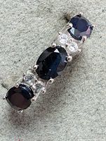 Close-up top view of ring showing the three dark blue sapphires separated by pairs of clear round accent stones set in sterling silver.