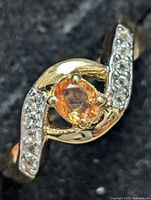 Close-up of sterling silver ring featuring central oval orange sapphire and surrounding white topaz stones on black background.