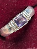 Close-up photo of sterling silver ring with square amethyst stone on red fabric background showing top and side details of setting and band.