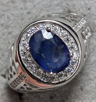 Close-up angled photo showing the blue oval sapphire center with surrounding clear stones set in sterling silver band with decorative cutouts on shoulders.