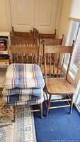 Six wooden antique chairs in natural finish with pressed backs, turned spindles, and spool stretchers. Five plaid seat cushions stacked on one chair.