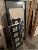 Various framed art pieces stacked leaning against wall, showing a large black framed floral artwork and three silver tone frames with multiple Asian prints.