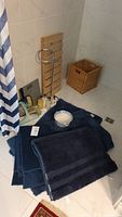 View of bath mats stacked on floor in bathroom, with chamber pot, wicker basket, wooden stand, toilet paper holder and toiletries visible.