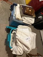 Two plastic tubs filled with folded white and off-white vintage bed linens, along with a book and a small basket on top of one tub.