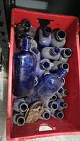 Red crate filled with numerous vintage cobalt blue glass bottles in various shapes and sizes, dusty, with an antique metal lock inside the crate