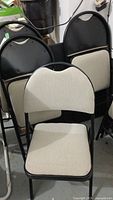 View of four black folding chairs with light beige upholstered seats and backs, stacked with one chair in front showing upholstery condition