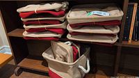 Stack of ten folded canvas and burlap tote bags with red bottom panel and white rope handles resting on wooden shelf.