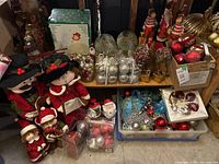Wide view of assorted Christmas decorations including cloth caroler dolls, packages of ornaments, small figurines and candle holders on shelving and floor