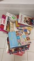 Pile of various crafting books with titles like Knitting, Quilted Dolls, Ultimate Visual Guide and others, plus container with decorative buttons.