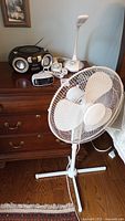 Photo showing all four items on and around a wooden dresser: the white floor fan standing on the floor, the Megatek portable CD player, the Ottlite desk lamp, and the small black Noma timer on top of the dresser.