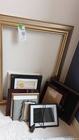 Assorted picture and painting frames standing against a wall; includes a large gold-toned frame and multiple smaller frames mostly backward showing backs and stand supports, and the digital photo frame with power cord.