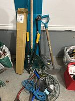 Full view of an assortment of outdoor gardening tools including hand saws with yellow handles, rakes, a shovel with blue handle, electric leaf blower, garden twine, gloves, and pruning shears.