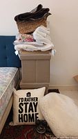 Stack of household items including throws, wicker basket with folded cloths, beige ottomans, decorative pillow, and black alarm clock on floor.