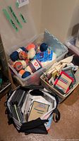 Plastic bins and bags holding assorted balls of yarn, knitting instructional books, DVDs and knitting needles.