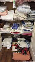 Photo of folded linens on closet shelves including white and pastel sheets, pillowcases, and multicolor blankets with fringe on the bottom shelf.