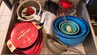 Drawer containing three red Sabatier melamine mixing bowls with spouts nested inside each other, blue nested plastic bowls, various glass measuring cups with red lids, and other kitchen utensils such as strainers and whisks.