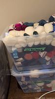 Two clear plastic totes stacked, each filled to capacity with yarn skeins in multiple colors and labeled “100% Wool/Natural.”