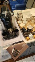 Nespresso Vertuo coffee machine with assorted cups and mugs on a tray, plus instruction booklet visible.
