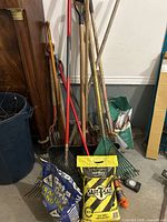 Full view of outdoor tools leaning against wall including rakes, shovels, pitchfork, broom; two bags on floor: blue Get a Grip Traction Sand and yellow Windsor Safe-T-Salt Ice Melt; coil of orange rope and timer device on floor.