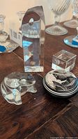 Three glass and crystal paperweights including a frog figurine, laser cut cube, and round faceted crystal piece on wooden surface.