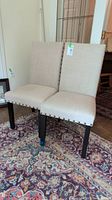 Two Parsons chairs placed side by side on a patterned oriental rug. Beige linen-like upholstery, nailhead trim along bottom edges and dark wooden legs shown.