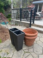 Photo showing two tall black wicker-style plastic planters next to one terra cotta-colored plastic planter on a stone patio.
