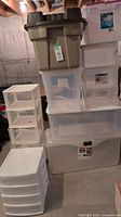 Stack of various plastic storage totes and three clear drawer units with labels visible, showing sizes and brands.