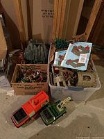 Overall view of the large Playmobil castle components packed in a plastic bin and cardboard box, two larger vehicles in front, on a concrete floor basement area.