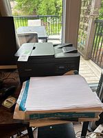Canon printer with paper stacked in front, placed beside a window with outdoor view.