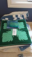 Four folded quilts displayed stacked, visible a blue and white quilt and a green quilt with large leaf pattern, along with two folded quilted pouches on top of the green quilt.