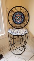 Photo showing the mosaic top plant table with black metal octagonal geometric frame, with the round metal framed wall clock resting on top.