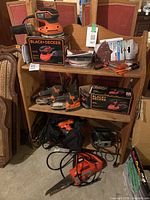Overall view of multiple Black+Decker tools and accessories on wood shelf in basement with chairs in background.
