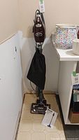 Full front view of Shark Rocket Deluxe Pro upright vacuum standing next to a shelf. Black fabric filter bag is attached. Instruction manual laying on floor in front.