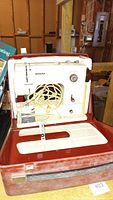 Photos of a cream-colored vintage Bernina Minimatic sewing machine with coiled power cord, presser foot attached, control dials, and red and white hard carry case showing wear and dust.