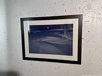 Framed digital photograph showing dark blue and black landscape with a small orange-yellow moon or sun, white mat and dark composite wood frame against a concrete wall.