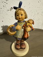 Front view of the Goebel figurine showing girl holding basket and doll, dressed in blue dress with blue bow in hair.