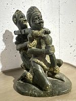 Side front angle of the green stone sculpture showing two kneeling African figures attached together with detailed golden accents and carved facial features.