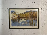 Framed watercolour artwork showing a landscape scene with water and trees mostly in blue, brown, and yellow tones.