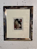 Full view of framed modern art with a silver metal frame and white mat, showing an abstract face design in muted tones.