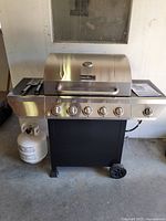 Full view of Nexgrill gas barbecue grill with stainless steel lid, six control knobs, black stand with wheels, propane tank attached on left side, and tools on left shelf.