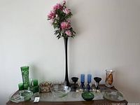 Full display of lot showing tall black vase with flowers in center, various coloured glass and crystal vases, glasses and bowls arranged on wooden table.