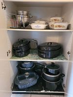 Full view inside cabinet showing an assortment of CorningWare glass casserole dishes with floral patterns and lids on the top shelf, stainless steel pots and strainers, Granite Ware pots, and black metal cookware with glass lids on lower shelves.