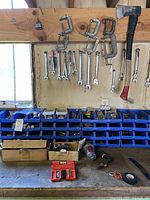 Workbench area showing hanging clasp clamps, crescent wrenches, combination wrenches, and organized blue bins with hardware below.